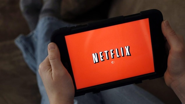 L'écran d'une tablette, tenue par un homme dont on aperçoit les pieds en arrière plan, affiche le logo de Netflix.
