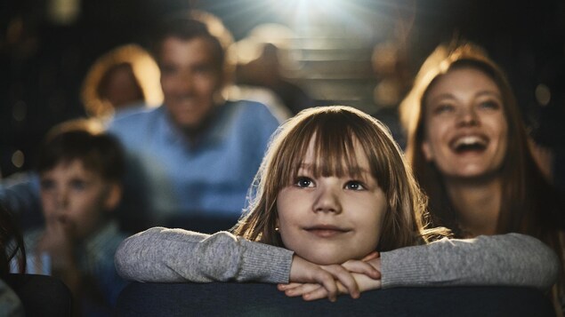 Une fillette appuie son menton sur ses mains, qui sont posées sur le dossier du siège devant elle, et regarde un film au cinéma avec un sourire.