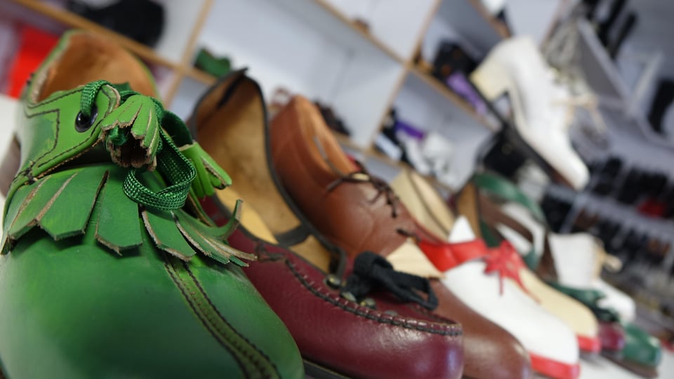 A row of leather shoes on a display shelf.  Some are green, red, brown and white.