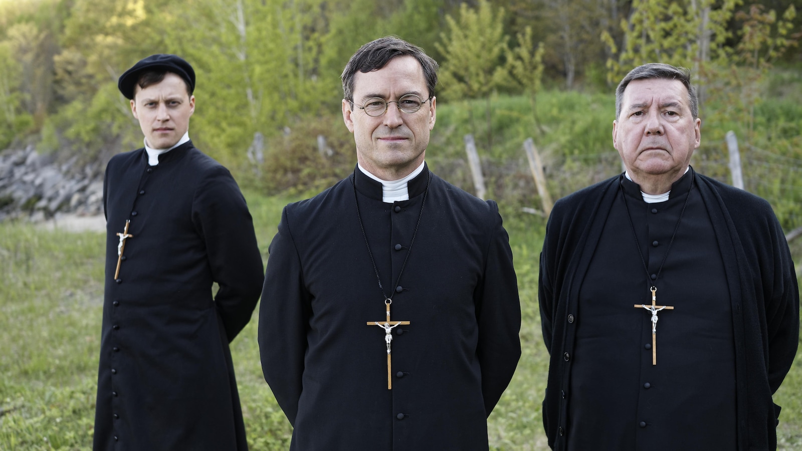 In a field, three men dressed as priests (including Sebastian Ricard and Remy Girard) look at the camera.