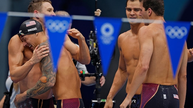 Swimmers hug each other to celebrate their victory.  A flag with the Olympic Games emblem flutters in the foreground. 