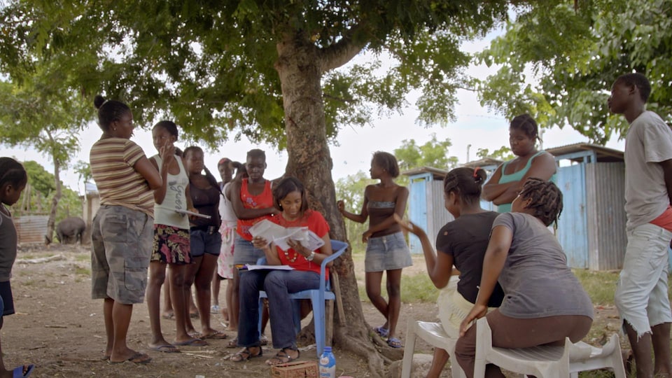 The woman is sitting in front of a tree and reading documents.  Men and women stand around.