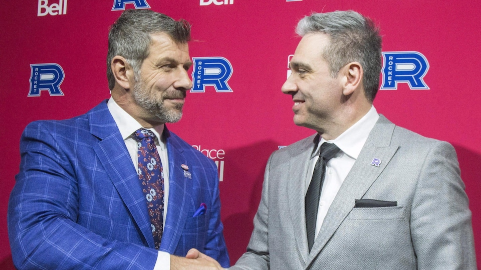 Two former hockey players, wearing jackets, shake hands and smile. 