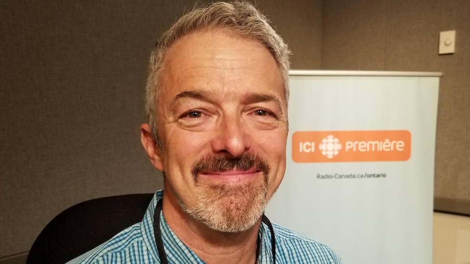 The individual is posed in front of an ICI Première sign in a radio studio.  He looks into the lens and smiles.