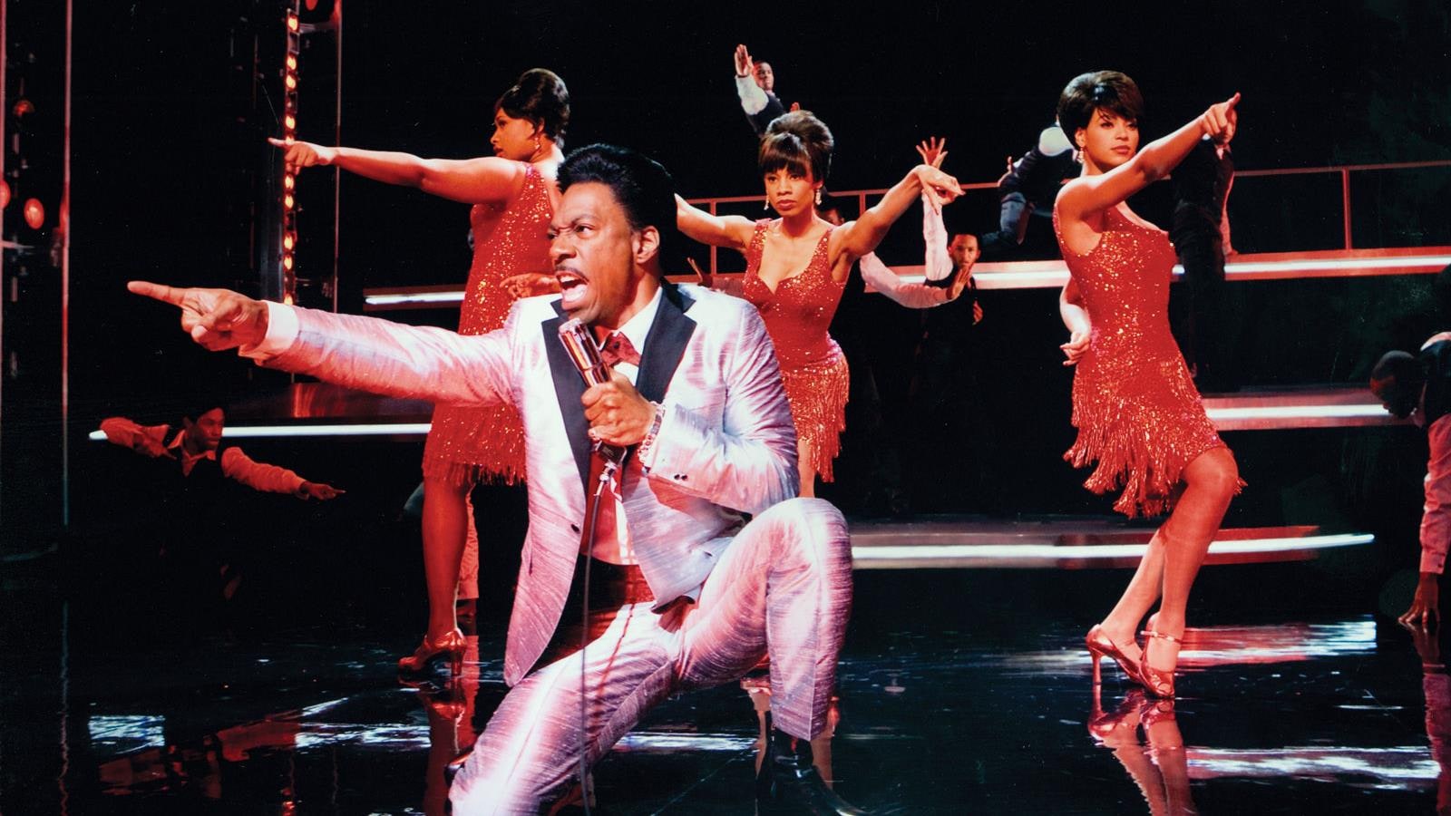 A man (Eddie Murphy) kneeling on stage singing in front of three girls in red dresses.