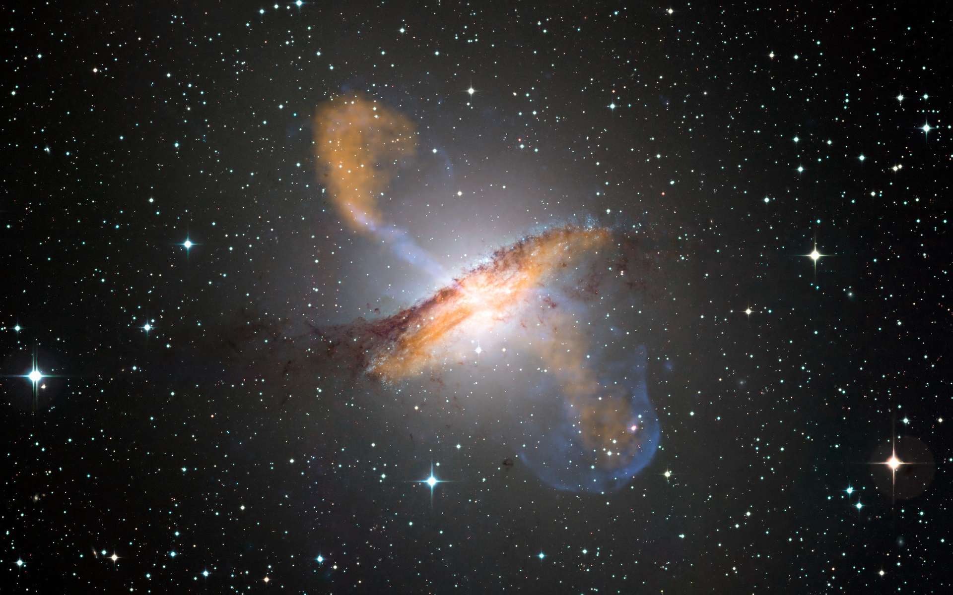 Image composite couleur de Centaurus A, révélant les lobes et les jets émanant du trou noir central de la galaxie active. Il s'agit d'un composite d'images obtenues avec trois instruments, fonctionnant à des longueurs d'onde très différentes. Les données submillimétriques à 870 microns, de Laboca sur Apex, sont indiquées en orange. Les données de rayons X de l'observatoire de rayons X Chandra sont indiquées en bleu. Les données de lumière visible de l'imageur à champ large (WFI) sur le télescope MPG/ESO de 2,2 m situé à La Silla, au Chili, montrent les étoiles et la bande de poussière caractéristique de la galaxie dans des couleurs proches de la « vraie couleur ». © ESO/WFI (optique) ; MPIfR/ESO/Apex/A.Weiss et al., (submillimétrique) ; Nasa/CXC/CfA/R.Kraft et al., (rayons X)