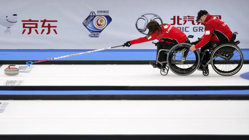 Curler Ina Forrest throws a stone with the help of teammate Dennis Thiessen, who is clutching her wheelchair.