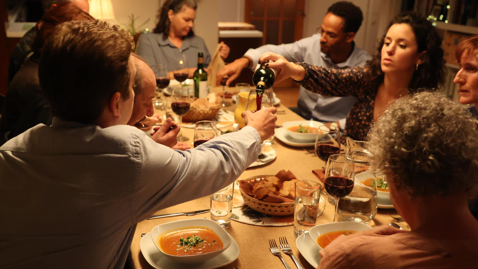 Several people share the meal around a table and one person serves red wine to one of the guests. 
