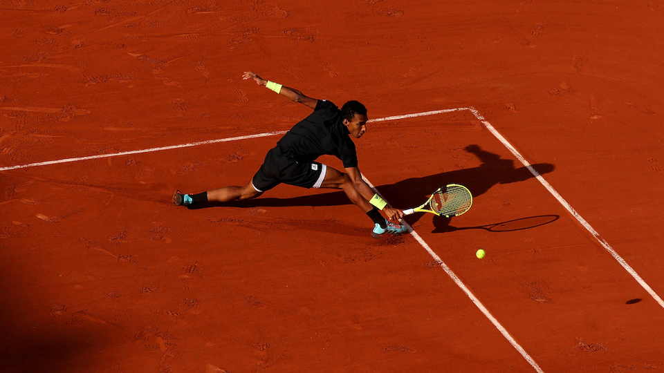 A tennis player in black stretches to hit a back ball with his yellow racket on a red clay court. 