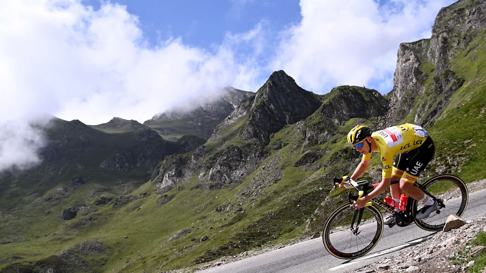 The cyclist, wearing the leader's yellow jersey, is on an incline.  Behind him we see large mountains. 