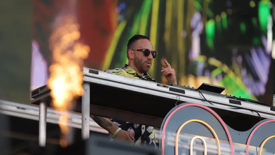 DJ behind his turntable.  He wears sunglasses and points to it with his left hand. 