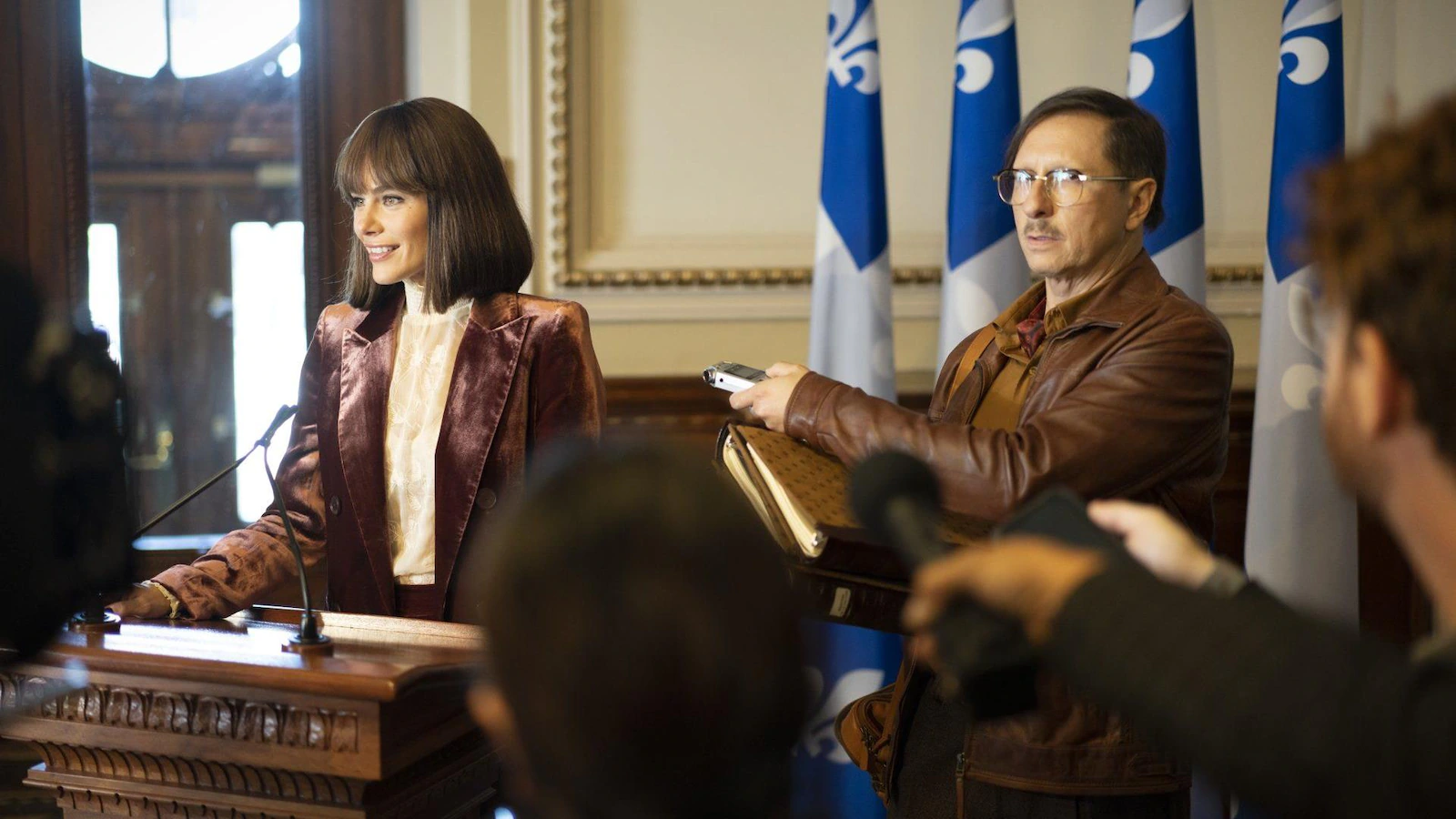 The woman speaks into a microphone behind a podium in a place reminiscent of the National Assembly Press Room, and behind them are Quebec flags.  A man holds a tape recorder next to her.
