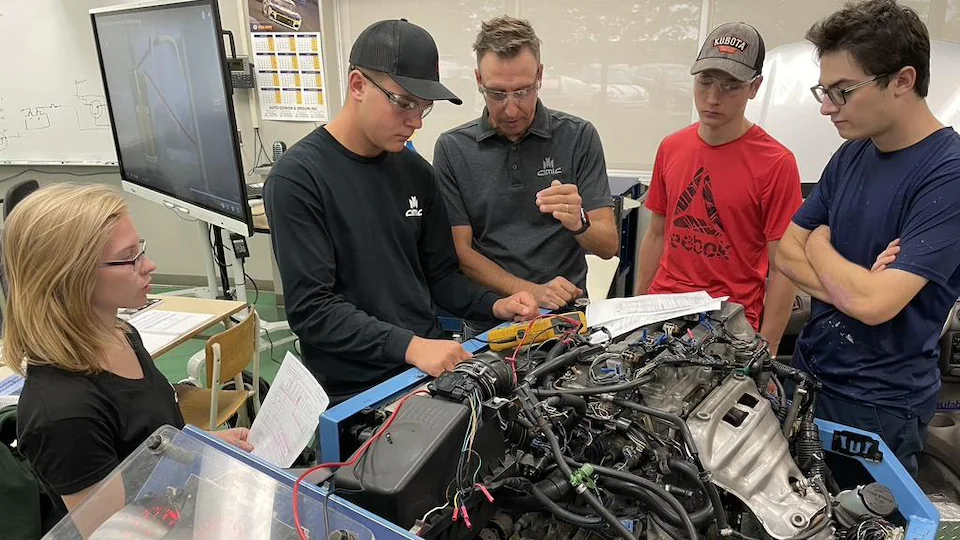 A car mechanic teacher in front of the engine talking to four attentive students.